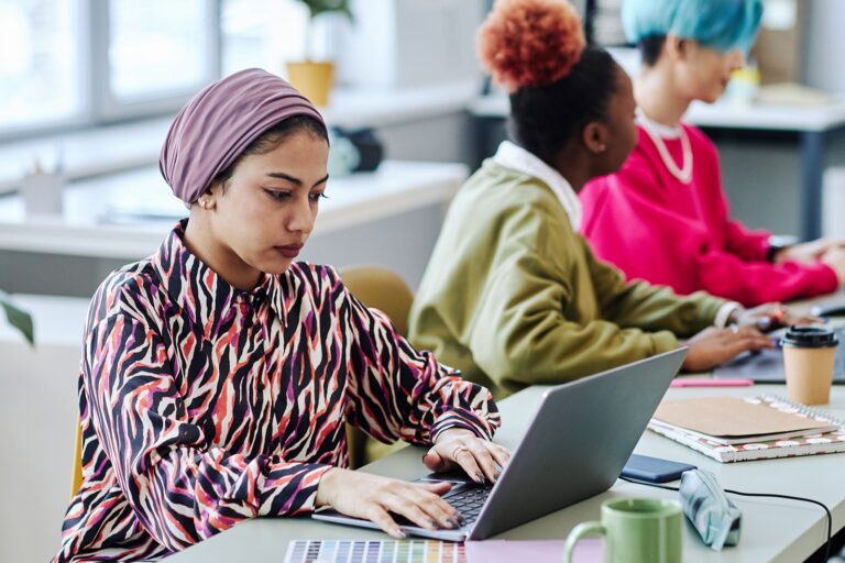 Three women sitting at a long desk working on their laptops. Two of the three women are collaborating.