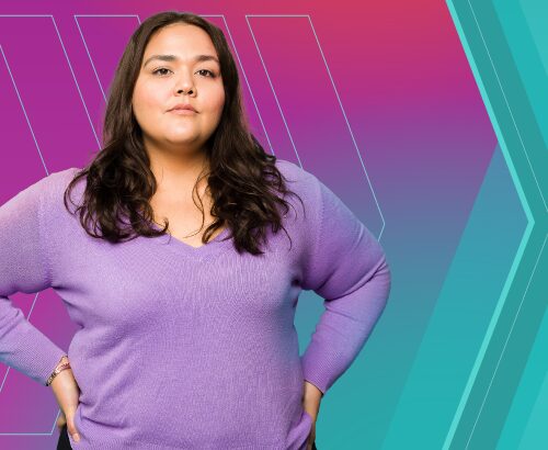 Light-skinned woman with brown hair wearing a purple, long-sleeved shirt, standing with her hands on her hips.
